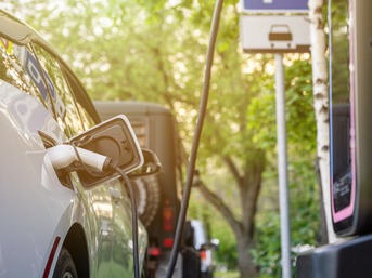Ein Elektroauto wird an einer öffentlichen Ladesäule geladen; weitere Fahrzeuge stehen dahinter in einer grünen, baumgesäumten Straße.
