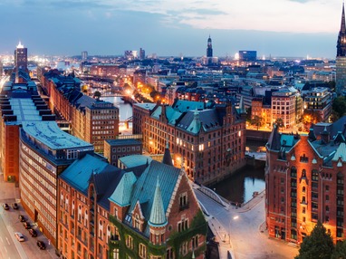 Abendliche Panoramaaufnahme der Hamburger Speicherstadt mit ihren historischen Backsteingebäuden, beleuchteten Brücken und Kanälen, sowie Blick auf die moderne Innenstadt und markante Kirchtürme im Hintergrund.