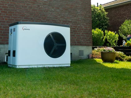 Auf dem Bild ist eine weiße solvis Wärmepumpe zu sehen. diese steht im garten, an der Rückwand eines Hauses. um die Wärmepumpe drum herum sieht man den Rasen und Pflanzen des Gartens. Die Wärmepumpe steht links im Garten und wird leicht seitlich betrachtet