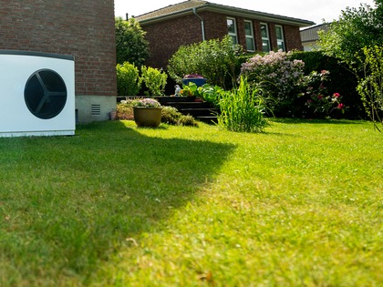 Auf dem Bild ist eine weiße solvis Wärmepumpe zu sehen. diese steht im garten, an der Rückwand eines Hauses. um die Wärmepumpe drum herum sieht man den Rasen und Pflanzen des Gartens. Die Wärmepumpe steht links im Garten und wird leicht seitlich betrachtet
