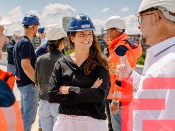 Mitarbeitende aus Netz- und Infrastrukturberufen stehen im Freien im Austausch; eine Frau mit Schutzhelm spricht lächelnd mit zwei Kollegen, alle tragen Sicherheitskleidung und Helme – Symbol für Zusammenarbeit und moderne Energienetz-Arbeit.