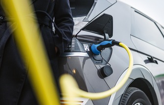 Nahaufnahme eines an die Ladebuchse angeschlossenen Ladegeräts für Elektrofahrzeuge, daneben eine Frau in einem schwarzen Mantel. Die Zukunft nachhaltiger Mobilität. Reduzierung des CO₂-Fußabdrucks.
