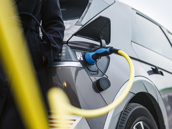 Nahaufnahme eines an die Ladebuchse angeschlossenen Ladegeräts für Elektrofahrzeuge, daneben eine Frau in einem schwarzen Mantel. Die Zukunft nachhaltiger Mobilität. Reduzierung des CO₂-Fußabdrucks.