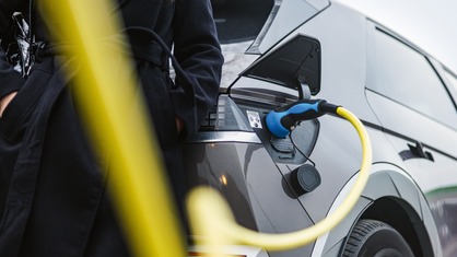 Nahaufnahme eines an die Ladebuchse angeschlossenen Ladegeräts für Elektrofahrzeuge, daneben eine Frau in einem schwarzen Mantel. Die Zukunft nachhaltiger Mobilität. Reduzierung des CO₂-Fußabdrucks.