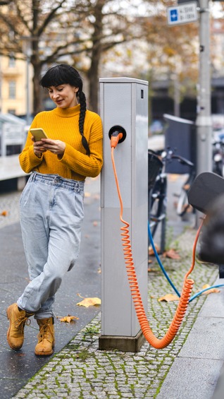 Junge Frau lehnt an einer Ladesäule und nutzt ihr Smartphone, während ihr Elektroauto lädt – Symbol für urbane E-Mobilität.