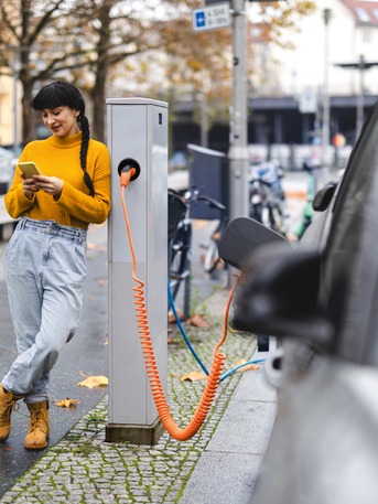 Junge Frau lehnt an einer Ladesäule und nutzt ihr Smartphone, während ihr Elektroauto lädt – Symbol für urbane E-Mobilität.