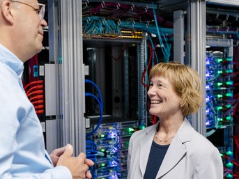 Zwei Personen führen ein Gespräch in einem Serverraum, umgeben von beleuchteten Serverschränken und zahlreichen Kabeln.