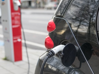 Ein in der Stadt parkendes E-Auto in der Nahaufnahme. Das Ladekabel ist am Auto eingesteckt. Im Hintergrund ist unscharf eine Ladesäule sichtbar.