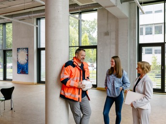 Ein Mann mit orangener Arbeitsjacke, Sicherheitshelm in den Händen, spricht mit zwei Frauen über die Arbeit.