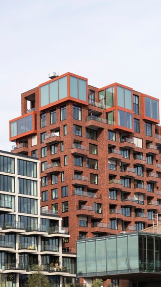Moderne Wohn- und Bürogebäude, darunter markante Fassaden aus Glas, Beton und rotem Backstein mit großen Fensterfronten und Balkonen, aufgenommen bei Tageslicht.
