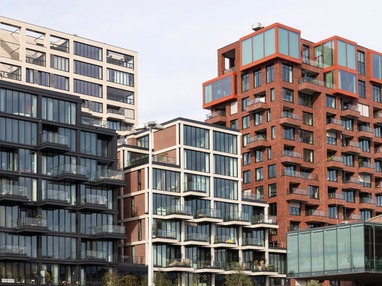 Moderne Wohn- und Bürogebäude, darunter markante Fassaden aus Glas, Beton und rotem Backstein mit großen Fensterfronten und Balkonen, aufgenommen bei Tageslicht.