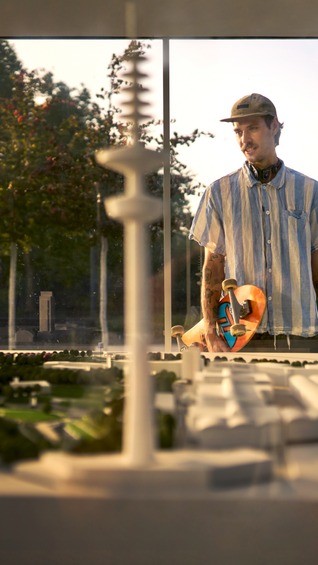Ein junger Mann mit Skateboard betrachtet durch eine Glasscheibe ein detailliertes Architekturmodell vom Hamburger Stadtpark im warmen Abendlicht; im Hintergrund stehen Bäume.