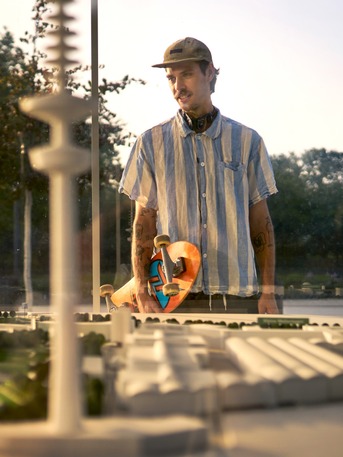 Ein junger Mann mit Skateboard betrachtet durch eine Glasscheibe ein detailliertes Architekturmodell vom Hamburger Stadtpark im warmen Abendlicht; im Hintergrund stehen Bäume.