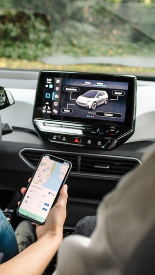 Innenraum eines Elektroautos mit zwei Personen; das Fahrzeugdisplay zeigt Fahrzeugdaten, während eine Person auf dem Smartphone eine Navigations- und Ladestations-App nutzt – Symbol für vernetzte E-Mobilität.