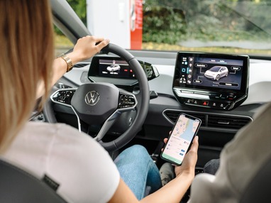 Innenraum eines Elektroautos mit zwei Personen; das Fahrzeugdisplay zeigt Fahrzeugdaten, während eine Person auf dem Smartphone eine Navigations- und Ladestations-App nutzt – Symbol für vernetzte E-Mobilität.