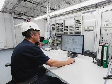 Mitarbeiter sitzt am Schreibtisch in der Leitwarte überwacht und steuert elektrische Anlagen über ein zentrales Leitsystem.